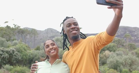 Diverse couple taking selfie on mountain trail, smiling on outdoor hike, scenic backdrop