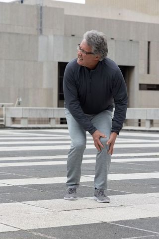 Senior man holding knee after outdoor exercise on urban rooftop plaza with striped tiles