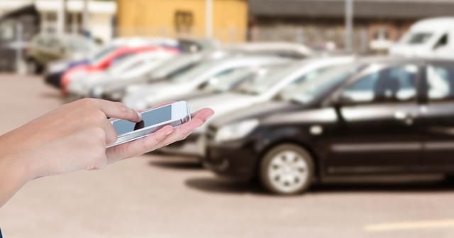 Hand holding smartphone with parked cars in background. Captures modern technology and communication. Ideal for use in articles about mobile apps, urban life, transportation solutions, and digital connectivity.