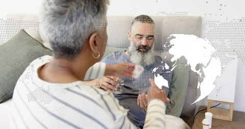 Caregiver handing medicine to bearded older man in bed with global health digital overlay