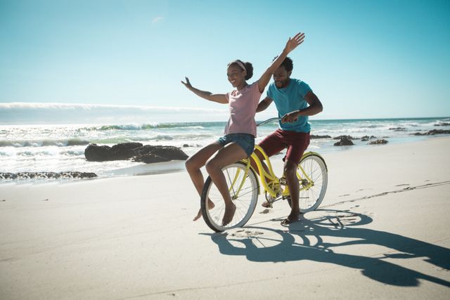 This image depicts a joyful African American couple riding a bicycle together along a sandy beach by the sea. The woman is sitting on the handlebars with her arms outstretched, while the man pedals behind her. The scene is bright and sunny, evoking feelings of fun, freedom, and romance. This image is perfect for use in travel brochures, romantic getaway promotions, lifestyle blogs, and advertisements focused on active and adventurous lifestyles.
