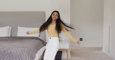 Young woman spinning in bright minimalist bedroom wearing mustard top and white pants, smiling