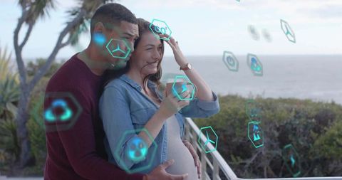 Couple embracing on ocean balcony touching pregnant belly with holographic ar health icons
