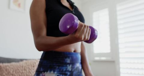 African American woman lifting purple dumbbell during home workout in bright living room