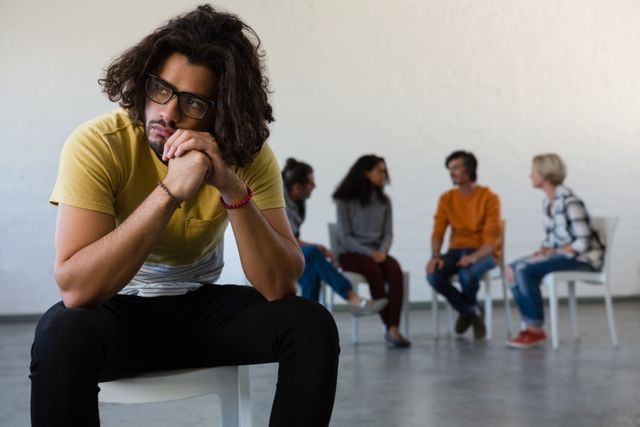 Man looking away while sitting on chair with friends discussing in background at art class