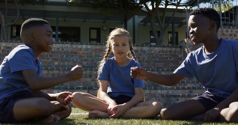 Diverse children playing hand game on school lawn, sitting cross-legged in blue shirts