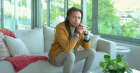 African american man with dreadlocks sitting on modern sofa in bright apartment interior