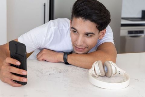Young man leaning on marble counter holding smartphone with white headphones nearby