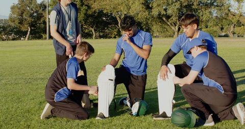 Diverse male cricket teammates preparing gear and adjusting pads on green field