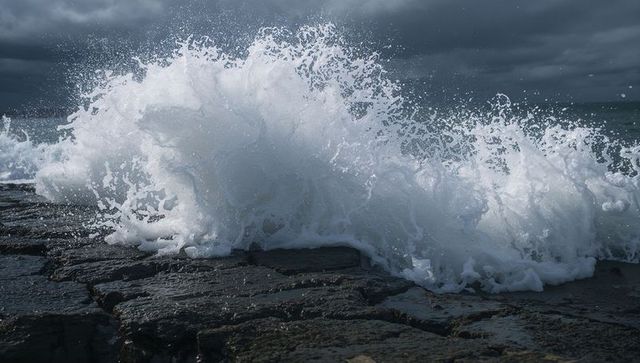 Powerful ocean wave crashing on dark basalt rock platform with foamy spray and stormy sky