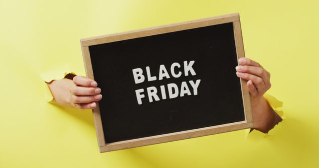Hands holding and pointing at black board with black friday sale text in white, on yellow background. Black friday, shopping, sale and retail concept digitally generated image.