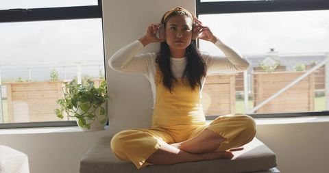 Asian woman in mustard overalls adjusting headphones while sitting by sunlit window