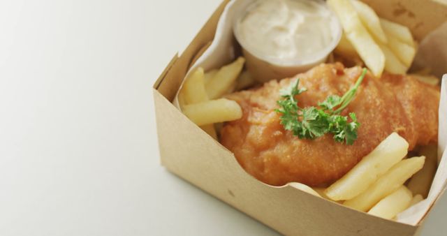 Fish and chips served in a takeout box with tartar sauce and parsley garnish. Ideal for illustrating fast food culture, British cuisine, or takeout meal concepts. Perfect for use in restaurant menus, food blogs, delivery services, or promotional materials.