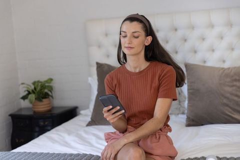 Calm woman relaxing on bed using smartphone in minimalist bedroom with terracotta accents