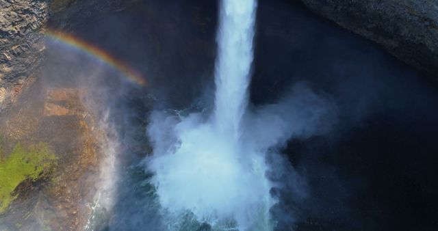 Aerial view of a powerful waterfall cascading into a pool below, with a faint rainbow visible in the mist. The force of the water creates a dynamic scene, emphasizing nature's power and beauty.