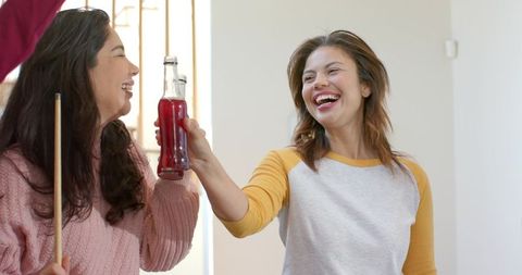 Friends laughing and clinking red soda bottle during casual sunlit home hangout