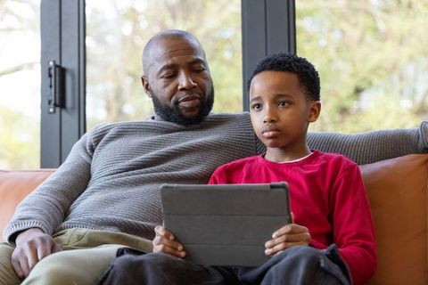 African american father and son sitting on sofa sharing tablet, family bonding at home