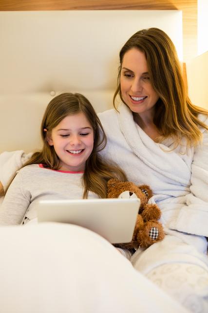 Mother and daughter relaxing together in bed, enjoying digital content on a tablet. Ideal for concepts like family bonding, modern parenting, technology use, leisure time, and home comfort.