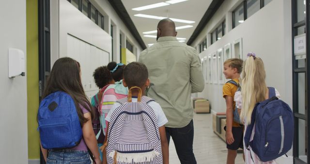Image shows teacher walking with group of diverse children in a school hallway. Suitable for content related to education, school environments, diversity in education, classroom management, academic settings, and student-teacher interaction visuals.