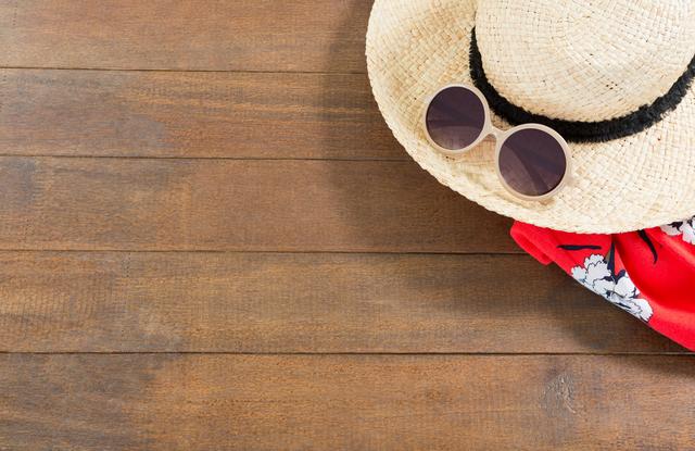 Wooden background displays summer beach accessories including a straw hat, sunglasses, and a red floral scarf. Ideal for depicting summer vacations, travel preparations, and seasonal promotions. Can be used in marketing materials for holiday destinations, travel agencies, or summer-themed advertisements.