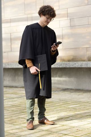Young graduate checking smartphone in cap and gown with yellow tassel on campus courtyard