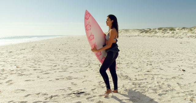 Woman in casual clothing holding a pink surfboard while standing on a sandy beach during a sunny day. This can be used for promotions concerning beach vacations, surfing lessons, summer activities, fitness and active lifestyle motivation, and travel blogs.