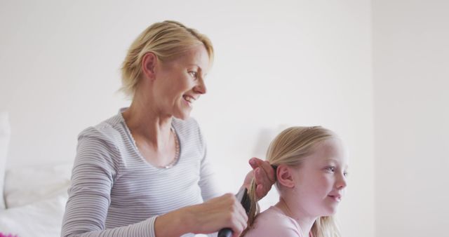 Happy caucasian mother brushing her daughter's hair and smiling in bedroom. Motherhood, childhood, domestic life, care and togetherness.