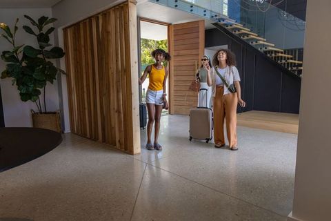 Diverse female travelers entering modern lobby with rolling suitcases, glass staircase, wood panel