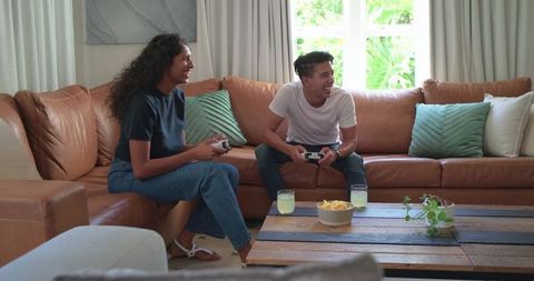 African american couple playing video games on sunlit living room sofa with snacks & plants