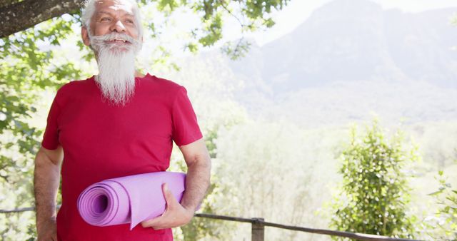 Elderly man holding a purple yoga mat while standing in an outdoor scenic area with trees and mountains in the background. Ideal for depicting healthy lifestyles for seniors, outdoor fitness, relaxation, or wellness programs for the elderly.