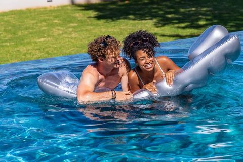 Young couple laughing on silver inflatable float in backyard pool on sunny summer day
