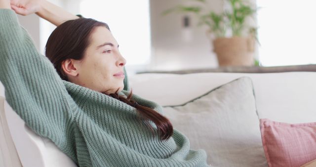 Woman sitting on couch in living room, appearing relaxed and content. Ideal for lifestyle, home comfort, and wellness themes. Suitable for blogs, articles, and advertisements focused on self-care, relaxation, and home living.