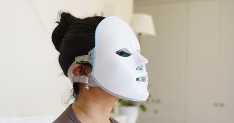 Woman wearing led facial therapy mask in bedroom for skin rejuvenation and relaxation