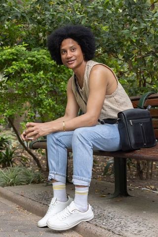 African man sitting on bench in urban park wearing casual outfit and carrying bag
