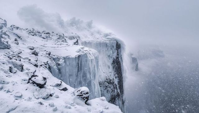 Frozen coastal cliff with icicle waterfalls, snowy plateau, blowing arctic snow and mist