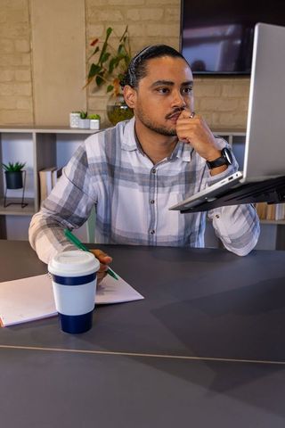 Man concentrating at desk while working remotely on laptop with notebook and coffee