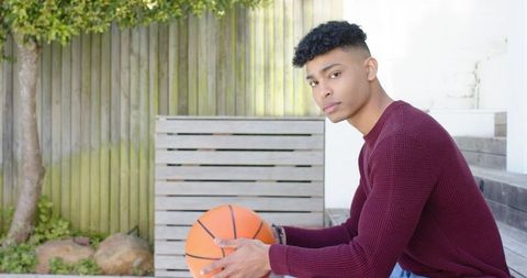Young man sitting on patio steps holding basketball in maroon sweater looking thoughtful