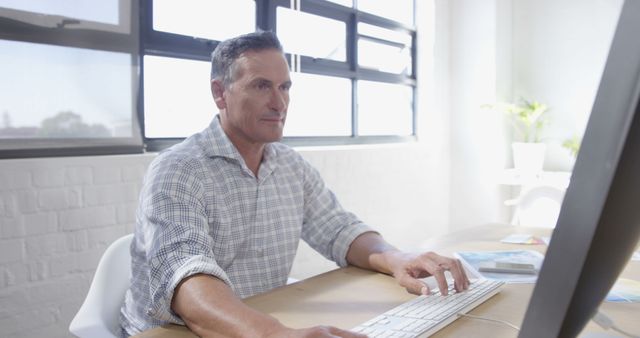 A middle-aged Caucasian man is focused on his work at a computer in a bright office environment, with copy space. His professional demeanor suggests he could be a businessman or an office worker managing daily tasks.