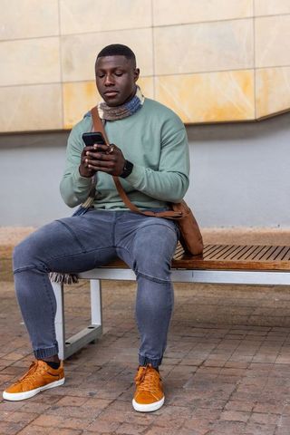 African american man checking smartphone on bench wearing scarf and orange sneakers