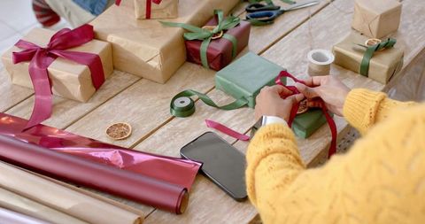 Tying red ribbon on green gift surrounded by kraft-wrapped presents and festive supplies