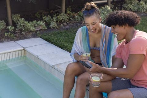 Young diverse couple laughing at smartphone poolside holding lemon drinks in swimwear