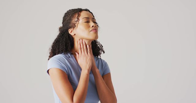 Young woman gently examining her thyroid gland as part of a self-assessment for potential health issues. She is standing against a neutral background. Suitable for articles or materials related to health assessments, medical self-care, thyroid conditions, and wellness education.
