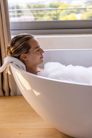 Woman reclining in bubble-filled freestanding bathtub by large window, serene morning light