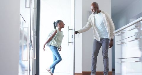 African american father welcoming teen daughter through modern glass door with backpack