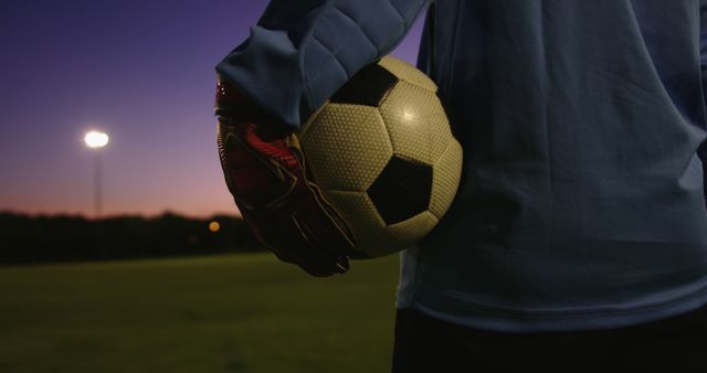 A soccer player is standing on a field at sunset holding a soccer ball. This is perfect for illustrating themes of sportsmanship, training sessions, soccer events, and athletic twilight scenes.