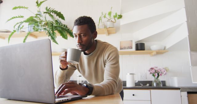 Happy african american man leaning on countertop in kitchen, using laptop and drinking coffee. Spending quality time at home alone.