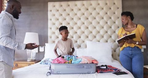 African american family packing suitcase on bed while mother writing travel checklist