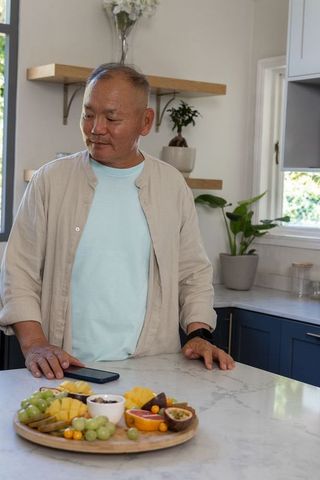 Mature asian man standing at marble kitchen island with fruit board and smartphone