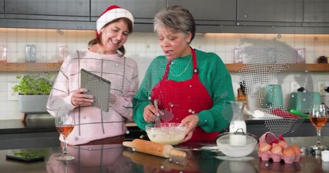 Middle-aged women baking holiday treats at kitchen island using tablet and santa hat