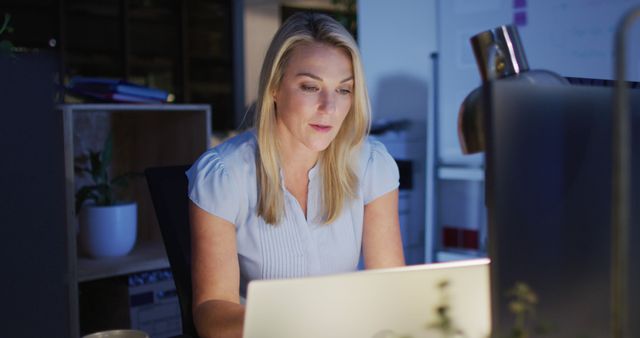 Woman working late in office, illuminated by laptop screen, conveys dedication and commitment to tasks. This can be used for articles or websites related to business, productivity, remote work, career dedication, and technology use in professional settings.